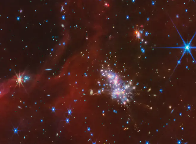 Aquí vemos la Nube de Digel S2. En el centro de la imagen hay un cúmulo de estrellas compacto compuesto por puntos luminosos de luz roja, azul y blanca. Débiles chorros con material grumoso y difuso se extienden en varias direcciones desde el cúmulo brillante. Arriba y a la derecha hay un cúmulo de estrellas más pequeño. Mechones de material rojo translúcido se extienden por la escena, aunque hay parches y un espacio notable en la esquina superior izquierda que revela el fondo negro del espacio. Las galaxias de fondo están dispersas a lo largo de esta franja de espacio, apareciendo como pequeños puntos azul-blanco y naranja-blanco o discos delgados y borrosos. Hay un punto azul-blanco notablemente más grande con picos de difracción, una estrella en primer plano en la esquina superior derecha. Crédito: NASA, ESA, CSA, STScI, M. Ressler (NASA-JPL)